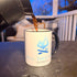 Coffee being poured into All Day Fire Outfitters mug outdoors. Campfire setting with ceramic mug for outdoor lifestyle.