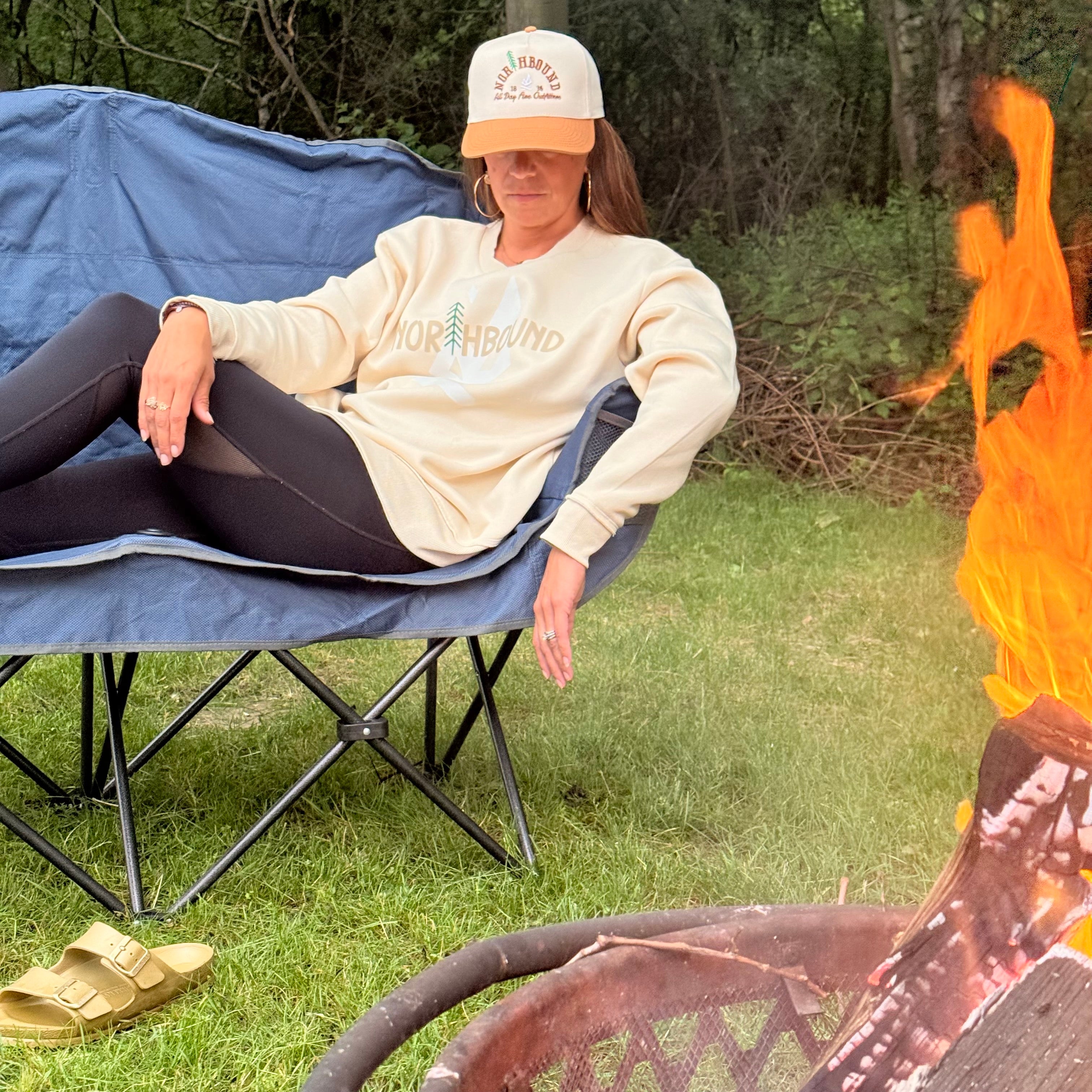 Woman lounging in a camping chair wearing the Northbound women's crewneck and Northbound snapback hat by All Day Fire Outfitters.